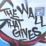The phrase "The Wall that Gives" is painted on a white wall