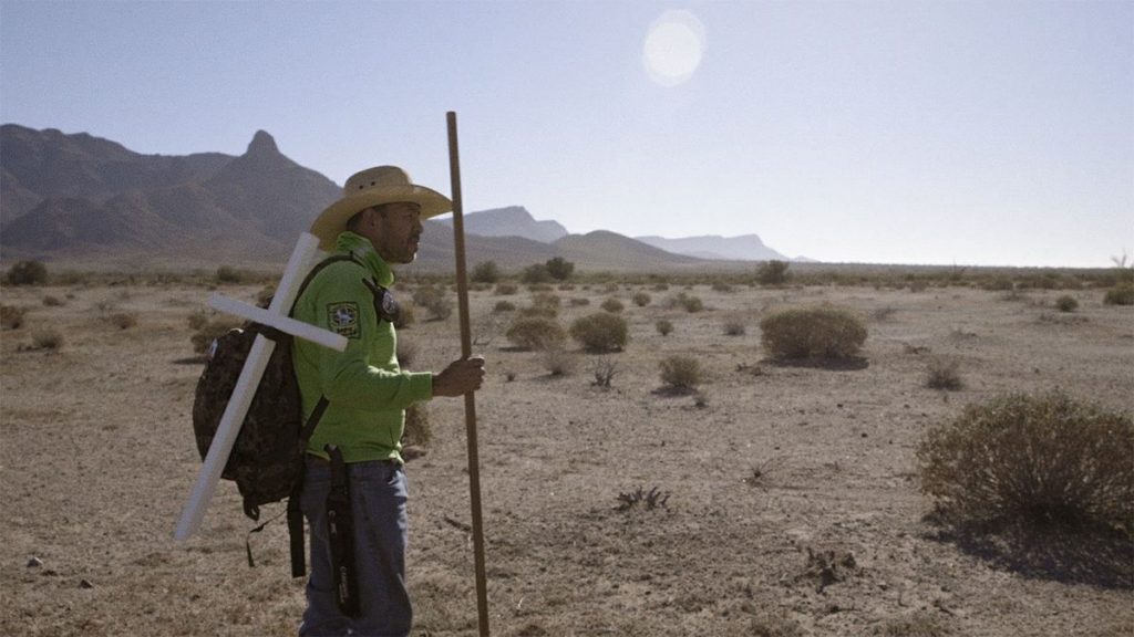 A person in the desert, holding a stick and wearing a hat, the sun behind them.
