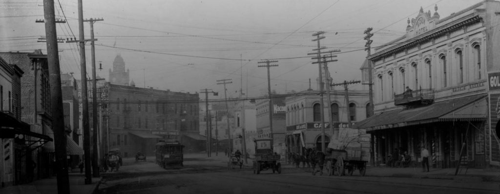 An image of the French district in downtown Los Angeles. The image shows Aliso Street in downtown Los Angeles, California, with signs labeling buildings "Griffins Transfer and Storage Co." and "Cafe des Alpes" next to "Eden Hotel," which are located on opposite corners of Aliso and Alameda Streets. A Pacific Electric streetcar sign reads "Sierra Madre" and automobiles and horse-drawn wagons are seen in the dirt road.