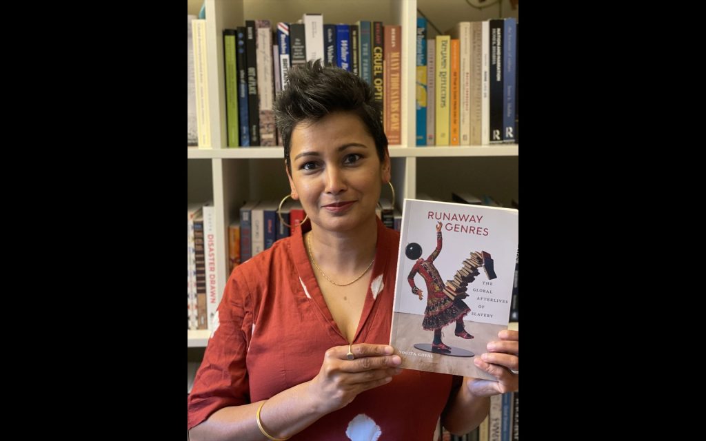 Yogita Goyal poses with her book against a bookshelf.