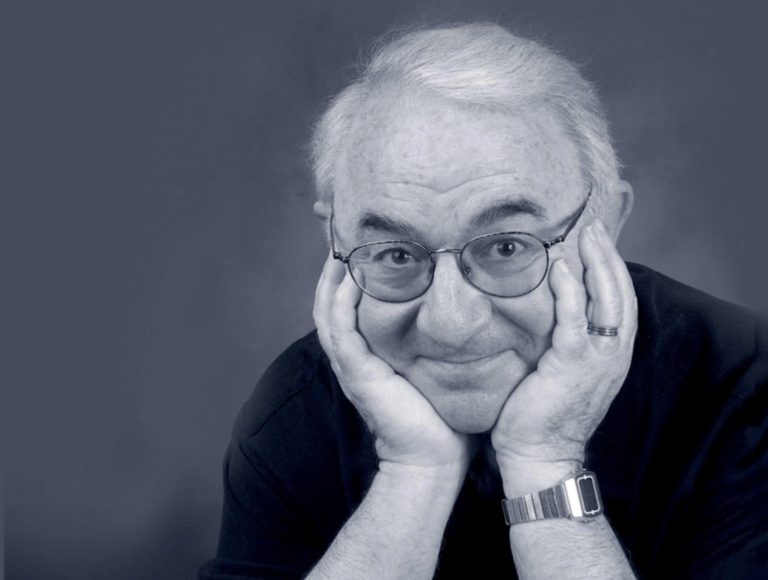 David Kaplan smiles mischievously with his cheeks in his palms in a black-and-white portrait