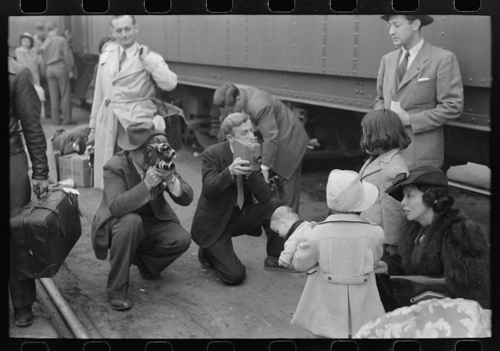 A black-and-white photo depicts cameramen snapping photos of a young Japanese girl as she waits to be sent from Los Angeles to the Manzanar prison camp.