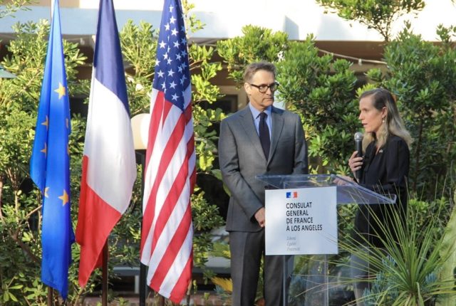 Dominic Thomas stands behind a lectern with another person, who is holding a microphone. The two are next to three flags, the European Union flag, the French flag, and the American flag.